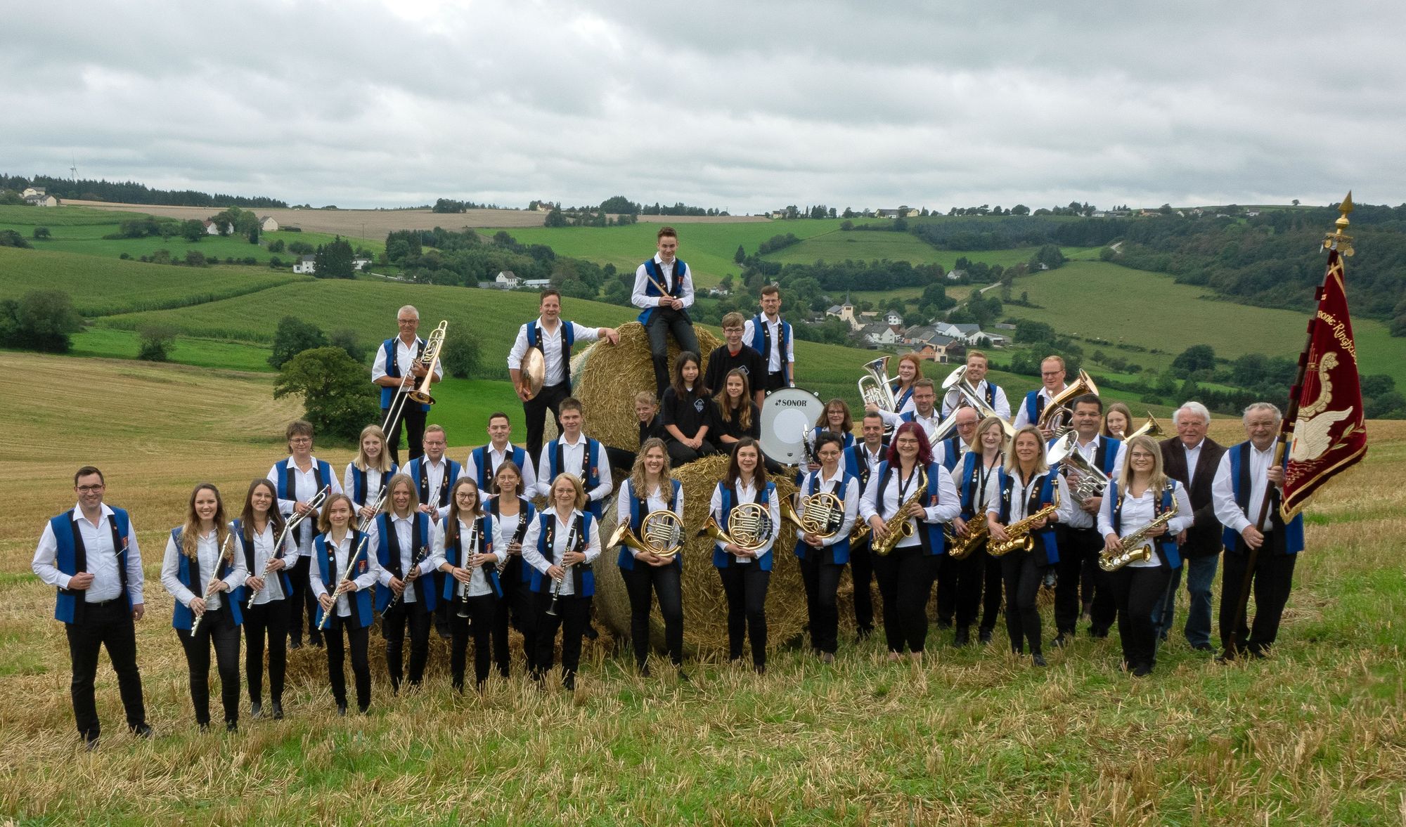 Mitglieder des MV "Harmonie" Ringhuscheid in Vereinstracht mit ihren Instrumenten auf einem Feld vor der Eifellandschaft